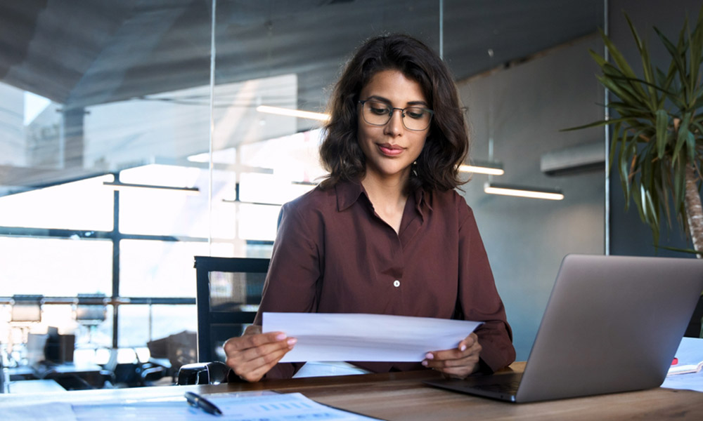 One woman with eyeglasses sitting at a desk in front of a silver laptop, holding a piece of white paper and looking down at it.