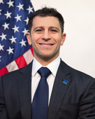 Headshot of Stephen Ehikian. Stephen wears a dark suit with a navy tie and a GSA lapel pin. He is seated in front of a U.S. flag