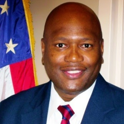 Portrait of Rob Miller, wearing a dark blue jacket, white shirt, and red and blue striped tie and is standing in front of an American flag