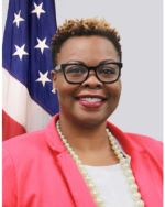  Smiling woman in a pink jacket and white top with a pearl necklace. A US flag is in the back left.