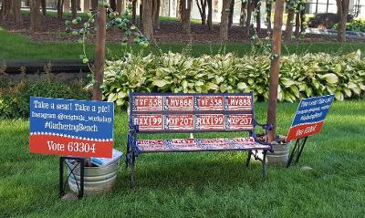 An artwork bench made of license plates and signs announcing it&#039;s title with trees in background