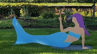 Painted aluminum sculpture of mermaid balancing a kinetic piece all in a park setting