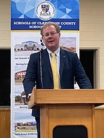 RA Jason Shelton speaking at podium in a blue suit and yellow tie 