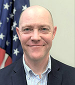 Portrait of David Rumsey, who is wearing a dark jacket and light blue shirt, and is standing in front of an American flag