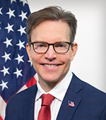 Headshot of Frank Schuler. Frank wears a blue plaid suit with a red tie.