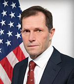 Headshot of Michael Peters. Michael wears a dark blue suit with a dark red tie and has a U.S. flag over his right shoulder.