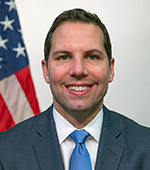 Portrait of Russ Riberto, who is wearing a blue jacket and tie, and standing next to the American flag