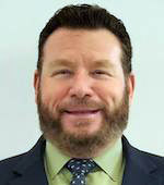 Portrait of Tom Lyman, who wears a dark jacket, light green shirt, and navy polka dot tie