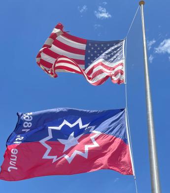 Two flag, American Flag on top, Juneteenth Flag on bottom flying on a flag pole with blue sky in the background