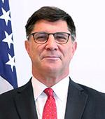 Portrait photograph of a smiling man with dark hair, glasses, black coat and red tie