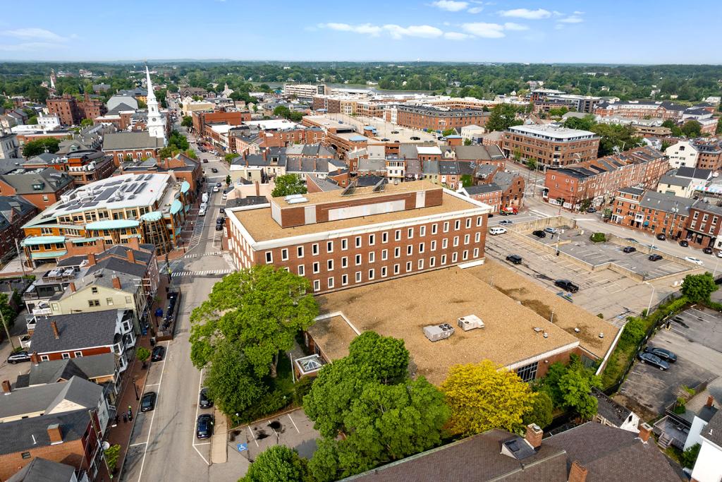 Aerial image of a four-story red brick building with a large one-story annex overlooking a two-level parking lot with loading docks and garage. The building sits in the middle of a downtown area with a white church and red brick buildings.