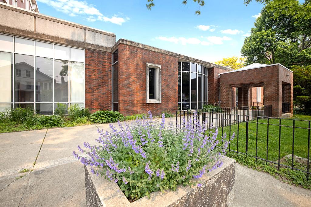 A sidewalk leads to a brick building with a large window and a portico with a skylight. Purple flowers grow from a planter. 