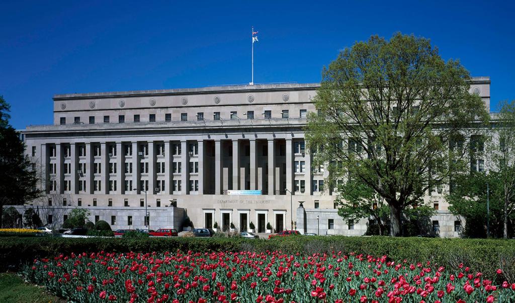 Light stone building with columns and flag pole, and green tree and bushes in front with many red tulips