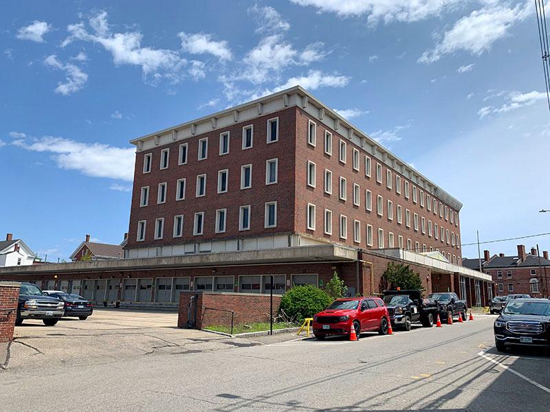 View of a 4-story red-brick building with a loading dock area and fenced parking lot