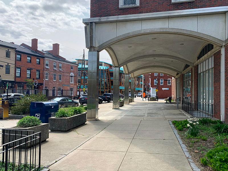 View of the portico and canopy of a red brick building with a sidewalk, planters, and neighboring businesses.