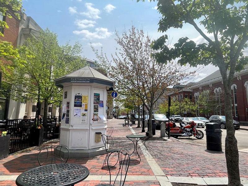 View of a brick sidewalk, a white kiosk, a street with parked cars, and several buildings