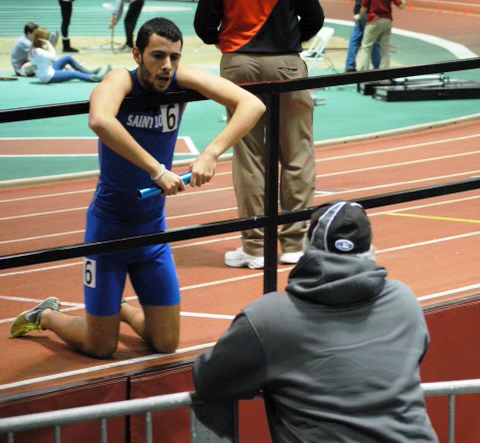 A medium-skinned teenage boy in a wrestling singlet kneels with arms hanging over a railing speaking to another man with his back to the camera. 