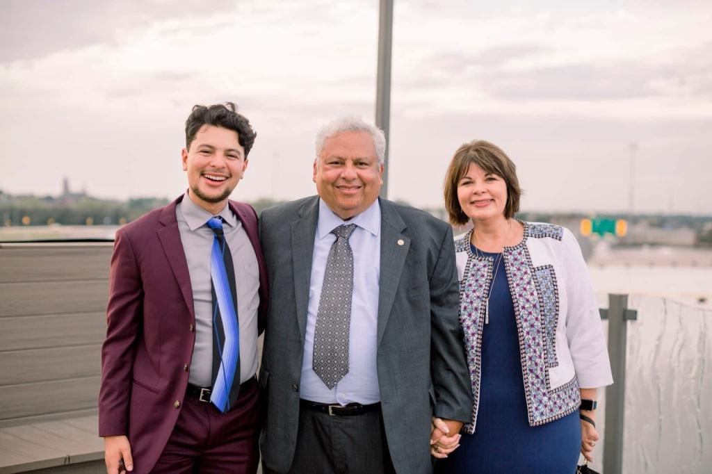 A medium-skinned, gray-haired man stands outdoors between a medium-skinned, dark-haired man and a light-skinned, dark-haired woman holding the woman's hand and putting his other arm around the man.