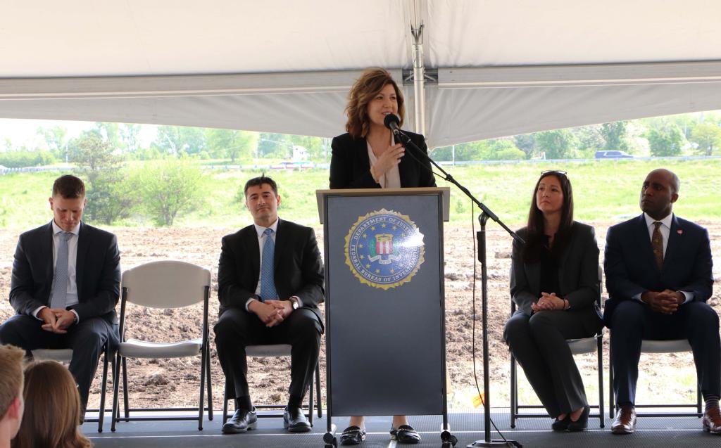 A person wearing business attire speaks into a microphone standing behind a podium while four other people behind them look on.