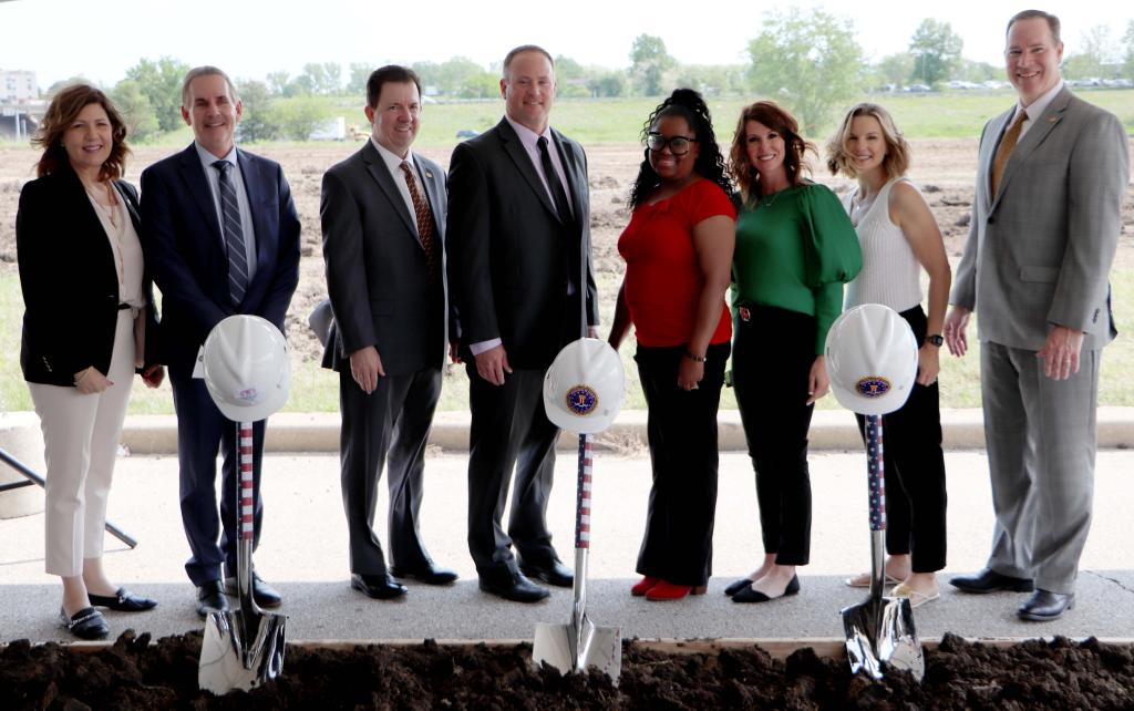 Eight people stand behind a box of dirt with three shovels stuck in it and hard hats on top of the shovel handles.