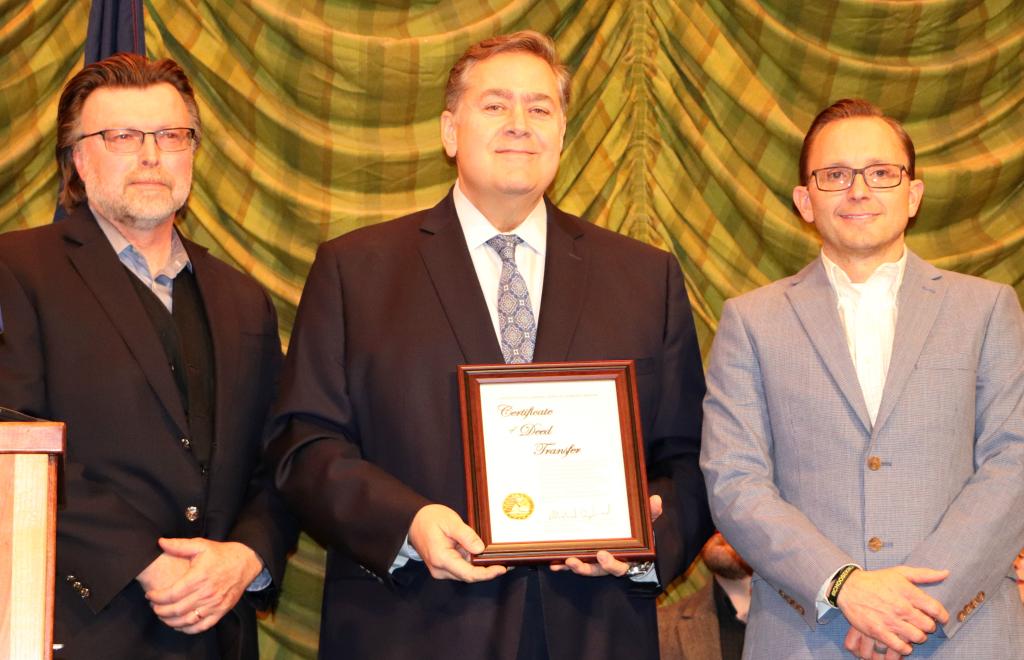 Three men stand in line horizontally and the center gentleman holds a framed Certificate of Deed Transfer