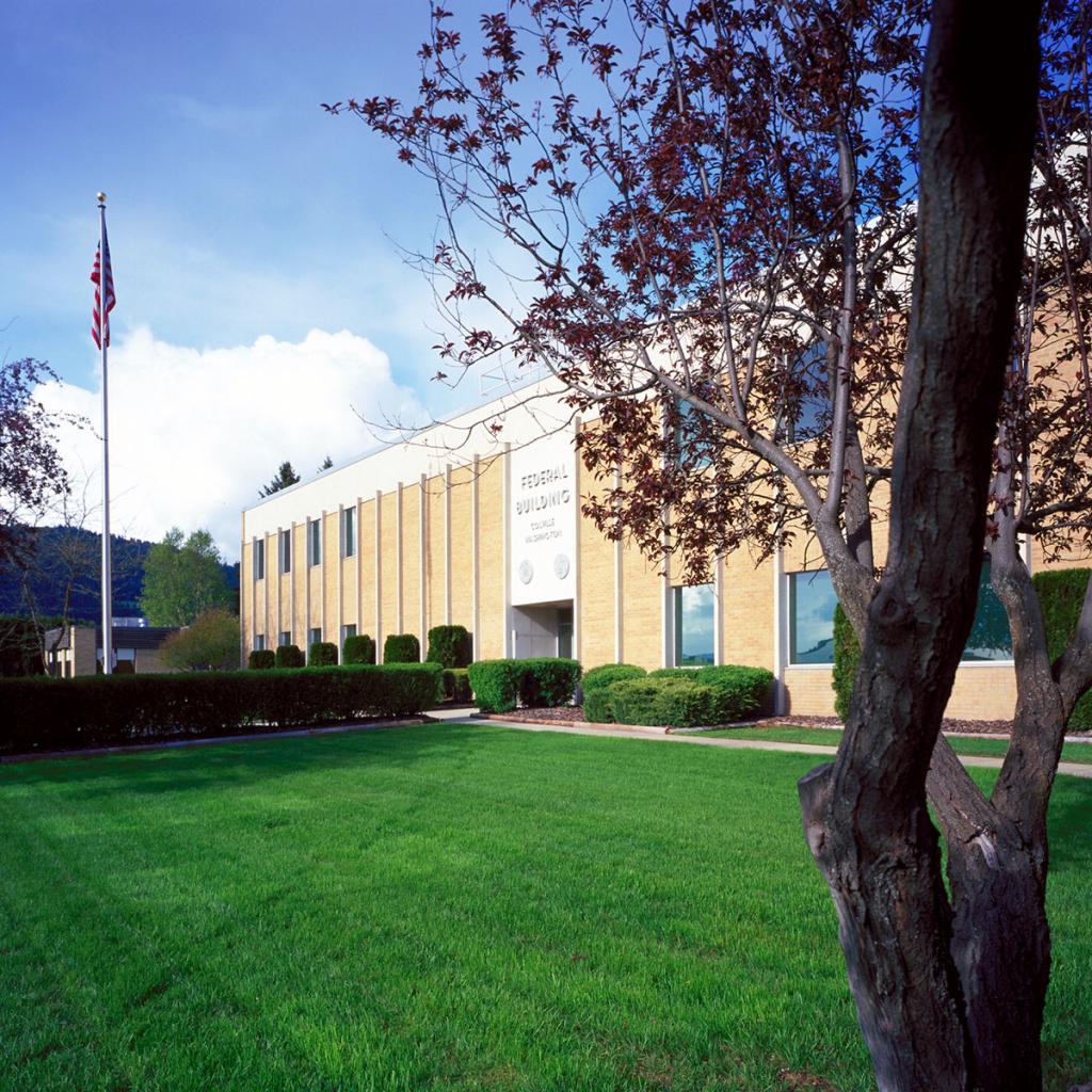 Angled view of two-level tan brick building with white sign above entrance, Federal Building, with hedges and green lawn and a U.S. flag on a pole and a red-leaf tree in the foreground on the right