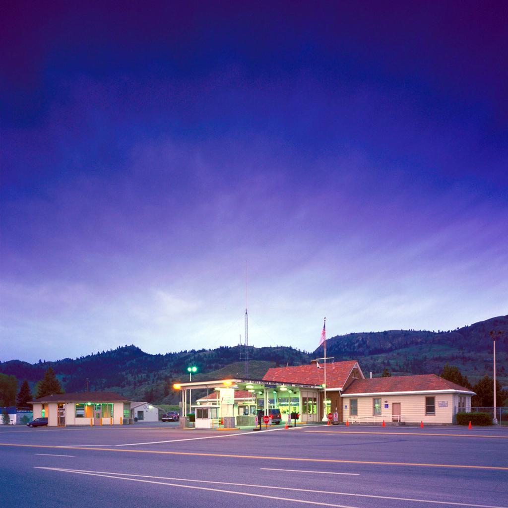 Tan and red low gable-roof buildings with canopy over entrance and two lanes in the larger building on the right, with pavement, low hills and vivid blue sky