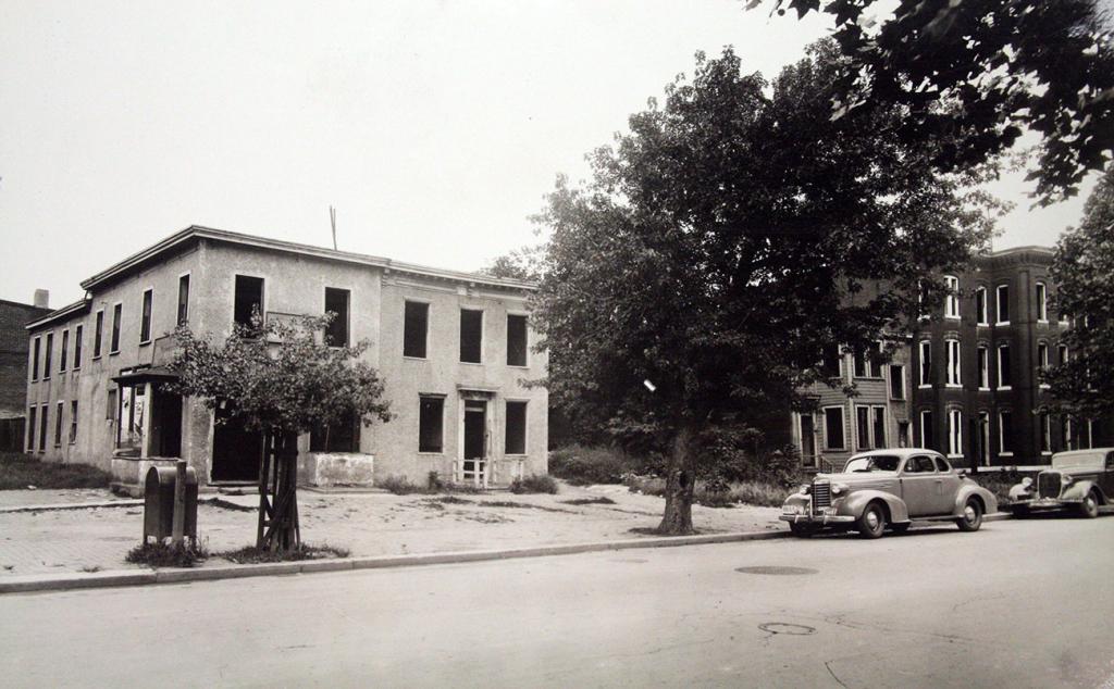 Black and white photo of a stone building on a street with a mailbox, trees and cars