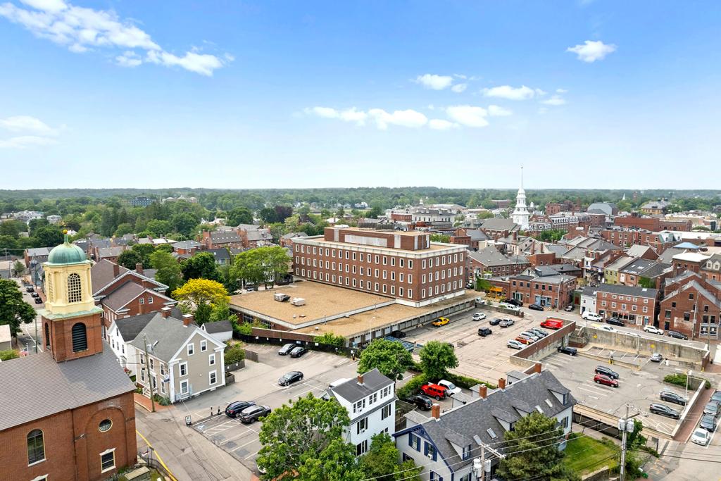 Aerial image of a four-story red brick building with a large one-story annex overlooking a parking lot and a downtown district with church spires.